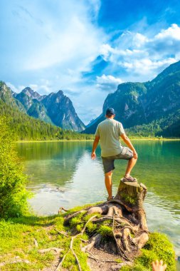 Bir ağaç kütüğünün üzerinde duran turist, yazın Dobbiaco Gölü 'nün ve Dolomite Dağları' nın büyüleyici manzarasına hayran kalıyor.