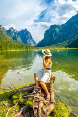 Hasır şapka takan genç bir kadın Dobbiaco Gölü kıyısındaki bir kütüğün üzerinde oturuyor. Dolomitlerin yaz gününde nefes kesen manzarasının tadını çıkarıyor.