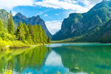 Büyüleyici alp manzarası Dobbiaco Gölü 'nün zümrüt sularını yansıtıyor, yemyeşil bitki örtüsü ve görkemli zirvelerle çevrili.