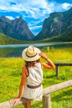 Hasır şapkalı bir kadın güneşli bir yaz gününde arka planında dolomitler olan Dobbiaco Gölü 'ne hayran.