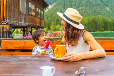 Anne ve oğlu göl kenarı aperatiflerinin tadını çıkarıyorlar. Renkli bir buzlu şeker ve serin bir dağ zeminine karşı sıcak bir içecek.
