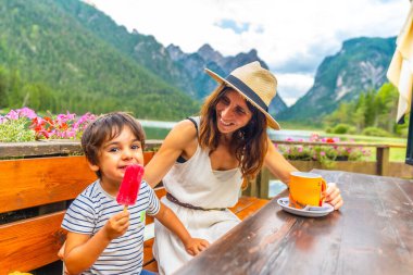 Anne ve oğlu göl kenarında çerezlerin keyfini çıkarıyorlar. Dolomitlerin içinde güzel bir dağ arkaplanı var.
