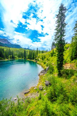Alp manzarası karersee gölünün turkuaz sularını yansıtıyor. Bulutlu mavi gökyüzünün altında, köknar ağaçları ve yeşil çayırlar ile çevrili.