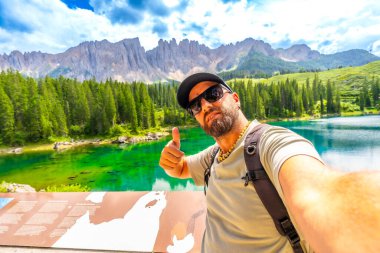 Turist, Carezza Gölü 'nün nefes kesici manzarasının tadını çıkarıyor. Dolomitler selfie çekiyor ve başparmaklarını kaldırıyor.