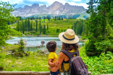 Anne oğlunu taşırken Carezza Gölü 'ndeki zümrüt sularının nefes kesici manzarasının tadını çıkarıyor. Dolomitlerle çevrili.