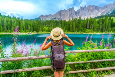 Sırt çantalı kadın yürüyüşçü, Carezza Gölü 'nün turkuaz sularına ve İtalyan dolomitlerindeki gecikmiş dağ sıralarına hayranlık duyuyor.