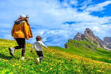 Çiçekli bir çayırda koşan turistler arka planda otlak ve seceda dağları, dolomitler, İtalya