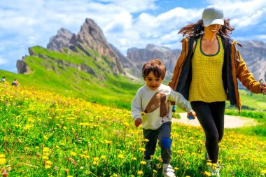İtalyan Alplerinde yaz tatilinin tadını çıkaran turistler, arka planda dağlarla birlikte kır çiçeklerinin olduğu bir tarlada koşuyorlar.