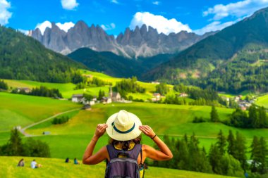 Bayan turist şapkasını tutarken arka planda küçük dağlarla birlikte Val di Funes 'deki St. Magdalena kilisesine bakıyor.