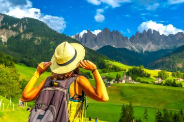 Şapkasını tutan kadın turist, Val di Funes 'daki Odin Dağları ve Santa Maddalena Kilisesi' nin çarpıcı manzarasına hayran kaldı.