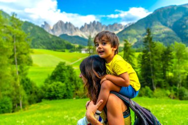 Mother carrying her son on her shoulders while enjoying the view of the odle mountains and the funes valley in the dolomites in summer clipart