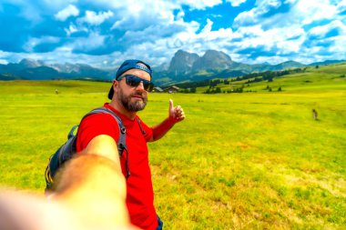 Sırt çantasıyla selfie çeken gezgin baş parmak işareti göstererek Dolomite, İtalya 'da Alpe di Siusi' de güzel bir yaz manzarasının tadını çıkarıyor.