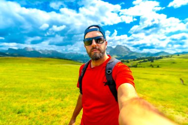 Güneş gözlüğü takan ve beysbol şapkalı bir turist güzel bir yaz gününde çekici dolomitlere karşı selfie çekiyor.
