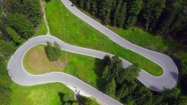 Dolomitlerdeki yemyeşil ormanlarla çevrili dolambaçlı bir dağ yolunda giden arabaların havadan görünüşü.