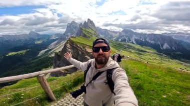 Sırt çantalı ve kameralı fotoğrafçı arka planda Di Eores dağ grubuyla birlikte uzanmış kollarıyla selfie çekiyor.