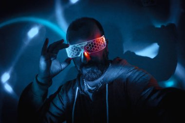 Sakallı adam, parlak parlak gözlükleri mavi ve turkuaz neon ışıklı siyah siber punk ayarıyla ayarlıyor.