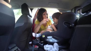 Bir kadın bir çocuğun araba koltuğuna oturmasına yardım ediyor. Çocuk sarı bir gömlek giyiyor. Kadın çocuğun elini tutuyor.