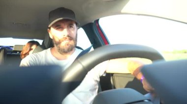 Bir adam arka koltukta bir kadınla araba kullanıyor. Şapka ve sakal takıyor.