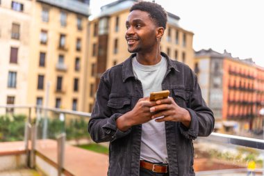 Afrikalı gülümseyen genç adam şehirde ayakta duran akıllı telefonu kullanarak bir mesaj gönderiyor.