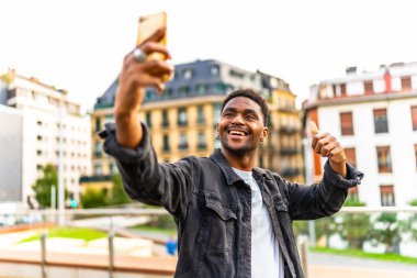 Kendine güvenen Afrikalı adam başparmağıyla hareket ediyor ve şehirde selfie çekiyor.