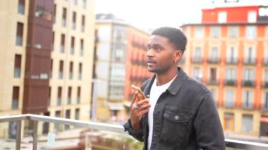 Afrikalı gülümseyen genç adam şehirde ayakta duran akıllı telefonu kullanarak bir mesaj gönderiyor.