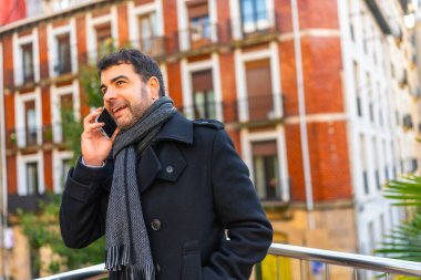 Zarif, kalın giysiler içindeki yetişkin beyaz adam şehirde ayakta duran cep telefonuyla konuşuyor.