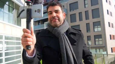 Beyaz erkek, zarif, olgun vlogger kullanıcısı, şehirdeki seyircilere bir şey açıklarken el sallıyor.