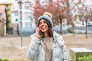 Happy and smiling caucasian young woman wearing winter clothes talking to the mobile in the city clipart