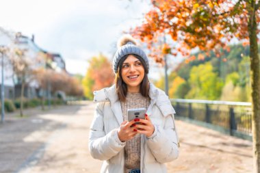 Bir kış günü şehirde cep telefonu kullanan mutlu, beyaz bir kadının ön fotoğrafı.
