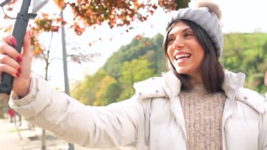 Beyaz tenli, genç ve güzel bir kadın vlogger. Sıcak kışlık giysiler giyip şehir parkında video kaydediyor.