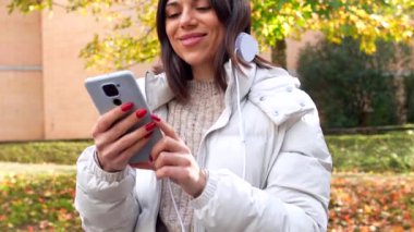 Sonbaharda şehir parkında duran cep telefonu ve kulaklıkla müzik dinlemeyi seven beyaz bir kadın.
