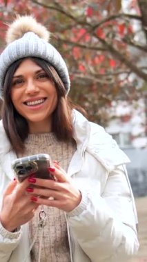 Mutlu ve gülümseyen beyaz kadın kışlık kıyafetler giyip şehirdeki cep telefonuyla konuşuyor.