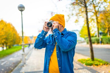 Kot elbiseli ve sarı yün şapkalı sıradan bir adam sokakta fotoğraf çekiyor.