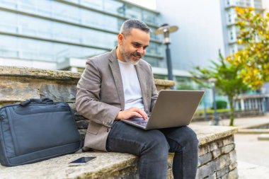 Finansal bölgede, taştan bir bankta oturup dizüstü bilgisayar kullanan mutlu beyaz iş adamı.