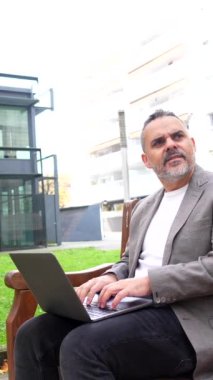 Girişimci dışarıda bir bankta oturuyor, bir dizüstü bilgisayarla çalışıyor, modern şehir ortamındaki görevlere odaklanıyor.