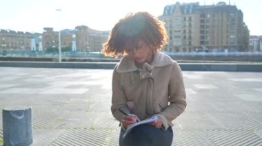 Kıvırcık saçlı güzel Latin kadın soğuk ve güneşli bir günde şehirde oturmuş notlar yazıyor.