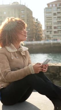 Nehrin kıyısında oturan Latin güzel kadın telefon kullanıyor.