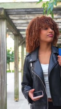 Deri ceket giyen genç Latin bir kadın elinde telefonla şehirde yürüyor.