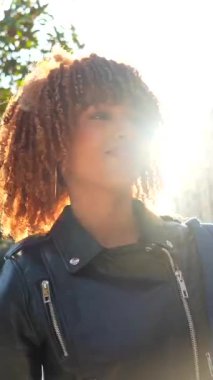 Kıvırcık saçlı, deri ceketli, gün batımında sokakta kameraya gülümseyen kendine güvenen genç Latin bir kadın.