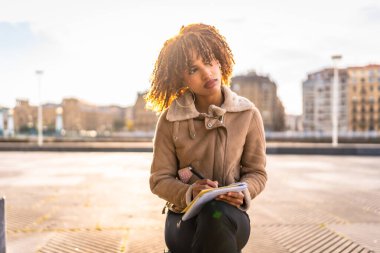 Düşünceli Latin kadın, soğuk bir günde arka planda şehir manzarası olan şehirde oturmuş notlar yazıyor.