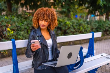 Parkta çalışan akıllı telefon ve dizüstü bilgisayar kullanan Latin güzeli genç bir kadın.