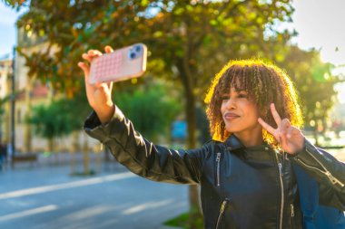 Kıvırcık saçlı güzel Latin kadın gün batımında şehirde selfie çekerken başarılı oluyor.