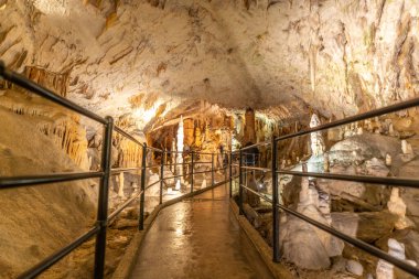 Turistler, Slovenia 'daki post-jna mağarasının içinde sarkıtlar ve dikitlerle çevrili aydınlık patikada yürümenin keyfini çıkarıyorlar.
