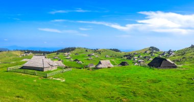 Dağılmış ahşap kulübeleri, canlı yeşil çayırları ve berrak mavi gökyüzünün altındaki yuvarlanan tepelerin manzarası.