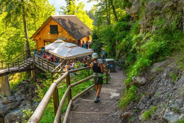 Kask takan turist Vintgar Vadisi 'nde yürüyor. Arka planda bir restoran dolusu insan var.