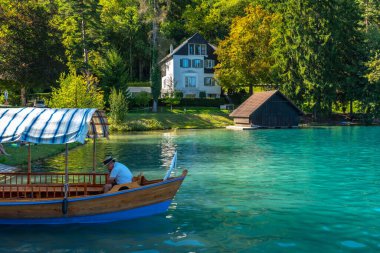Geleneksel pletna teknesi güzel gölde turistleri bekliyor. Arka planda bir villa ve kayıkhane var. Güneşli bir yaz gününde.