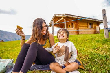 Mutlu anne ve oğul, yaz tatili boyunca dağlardaki ahşap bir kulübenin önünde battaniyede abur cubur yerken gülüyorlar.