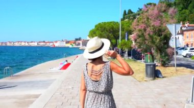 Hasır şapka takan genç bir kadın, yaz tatilinde, Piran gezinti alanında yürüyor, Adriyatik kıyılarının manzarasının keyfini çıkarıyor.