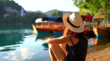 Yaz kıyafetleri içinde ve şapkalı genç bir kadın... Slovenia 'da kan kaybediyor. Geleneksel Pletna botlarının ve kilisenin manzarasının keyfini çıkarıyor.