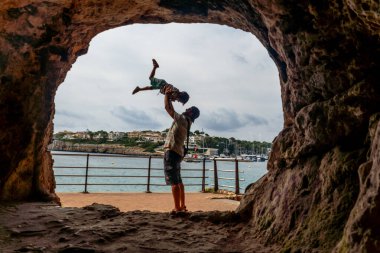 Baba, oğlunu neşe içinde kaldırıyor. Del Drac Mağarası ve Mallorca 'daki Akdeniz' in büyüleyici arka planına karşı.